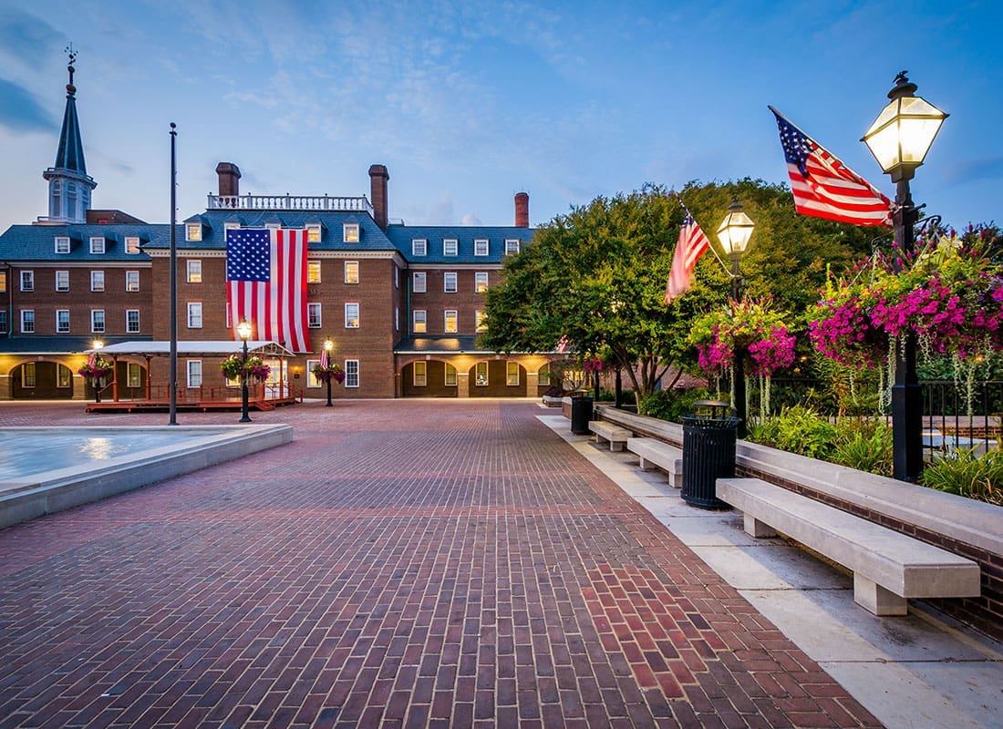 About Our Agency - Cobblestone Walkway in a Park with Lights American Flag and a Water Fountain in Historic Virginia with an Old Brick Building in the Background During the Evening