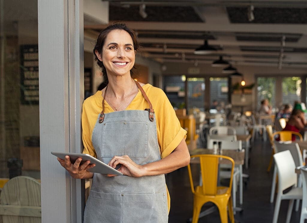 Business Insurance - Portrait of a Smiling Young Small Business Owner Wearing an Apron Standing by the Front Entrance of her Cafe While Holding a Tablet
