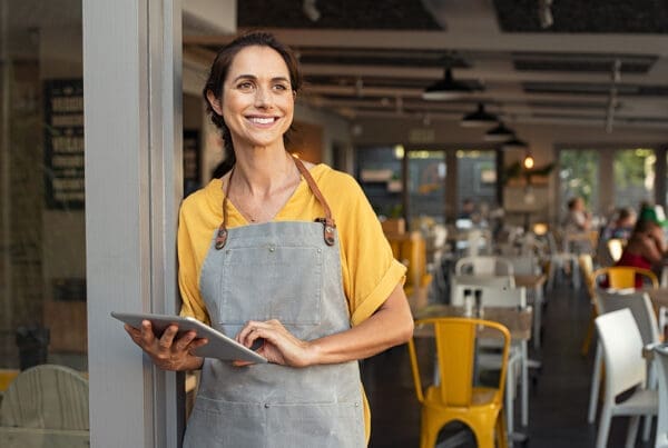 Business Insurance - Portrait of a Smiling Young Small Business Owner Wearing an Apron Standing by the Front Entrance of her Cafe While Holding a Tablet