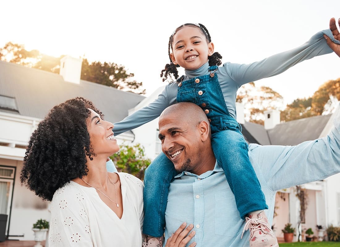 Personal Insurance - Cheerful Parents with the Father Giving his Young Daughter a Piggyback Ride Standing Outside Their Home on a Sunny Day at Sunset