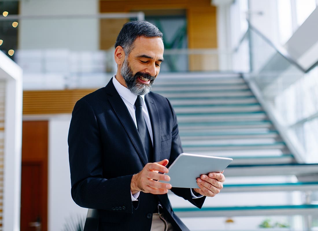 Blog - Middle Aged Businessman Standing in Front of a Staircase Inside a Modern Office While Looking at a Tablet he is Holding in his Hands